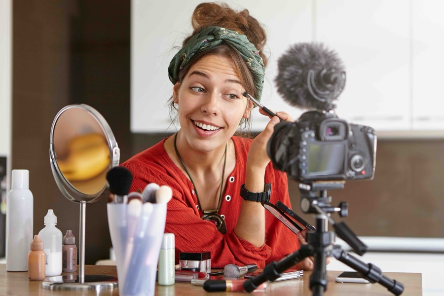 A beauty creator applying eye makeup to camera while filming on a tripod-mounted DSLR at a dressing table